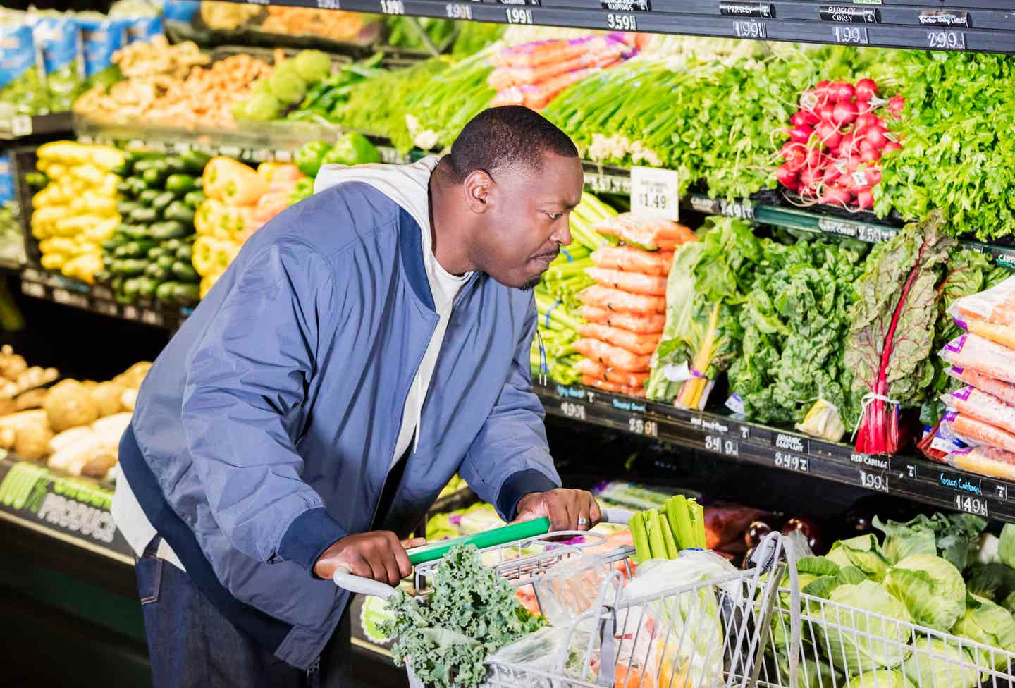 A Black man in his 40s shops the produce section of a grocery store, with a cart full of vegetables