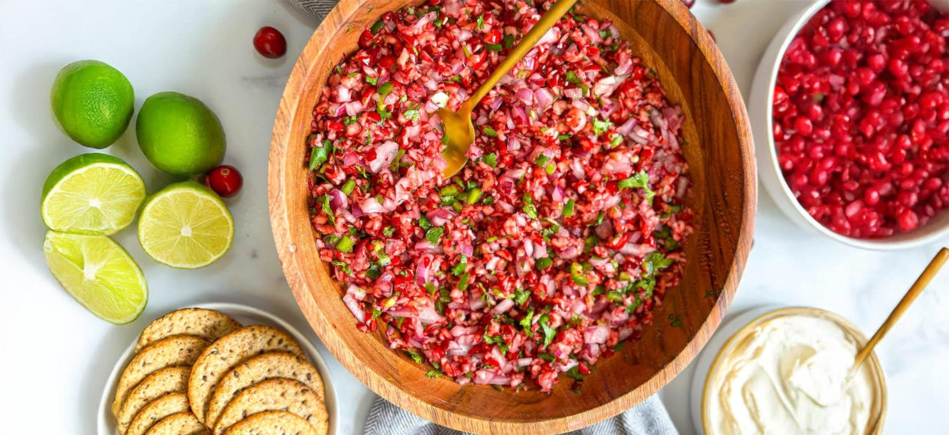 Jackie Akerberg’s Cranberry Salsa in a wooden serving bowl with a copper spoon, beside limes and crackers and vegan ricotta