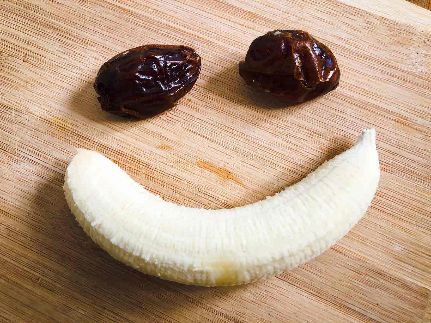 two dates above a peeled banana, forming a smiley face, on a wood surface