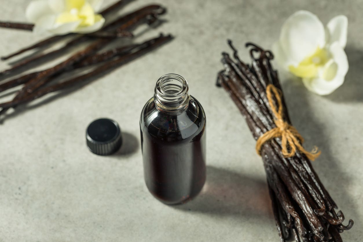 A bottle of vanilla extract beside whole vanilla bean pods and a flower