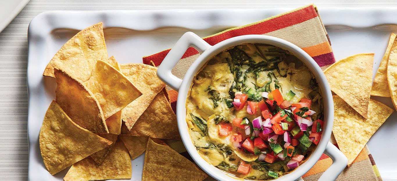 A small white ramekin filled with Cheesy Vegan Baked Spinach Dip sits atop a striped kitchen towel, with baked corn chips are scattered around