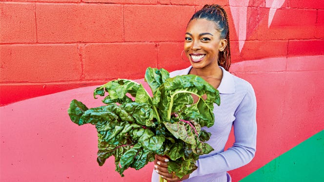 Cookbook author Jenne Claiborne holds a bushel of collard greens and smiles while standing in front of a bright pink and red wall outside.jpg