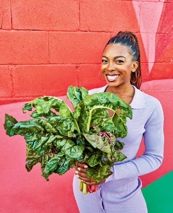 Author and vegan chef Jenné Claiborne of Sweet Potato Soul stands outside in front of a bright red wall, holding a large bushel of leafy greens and smiling