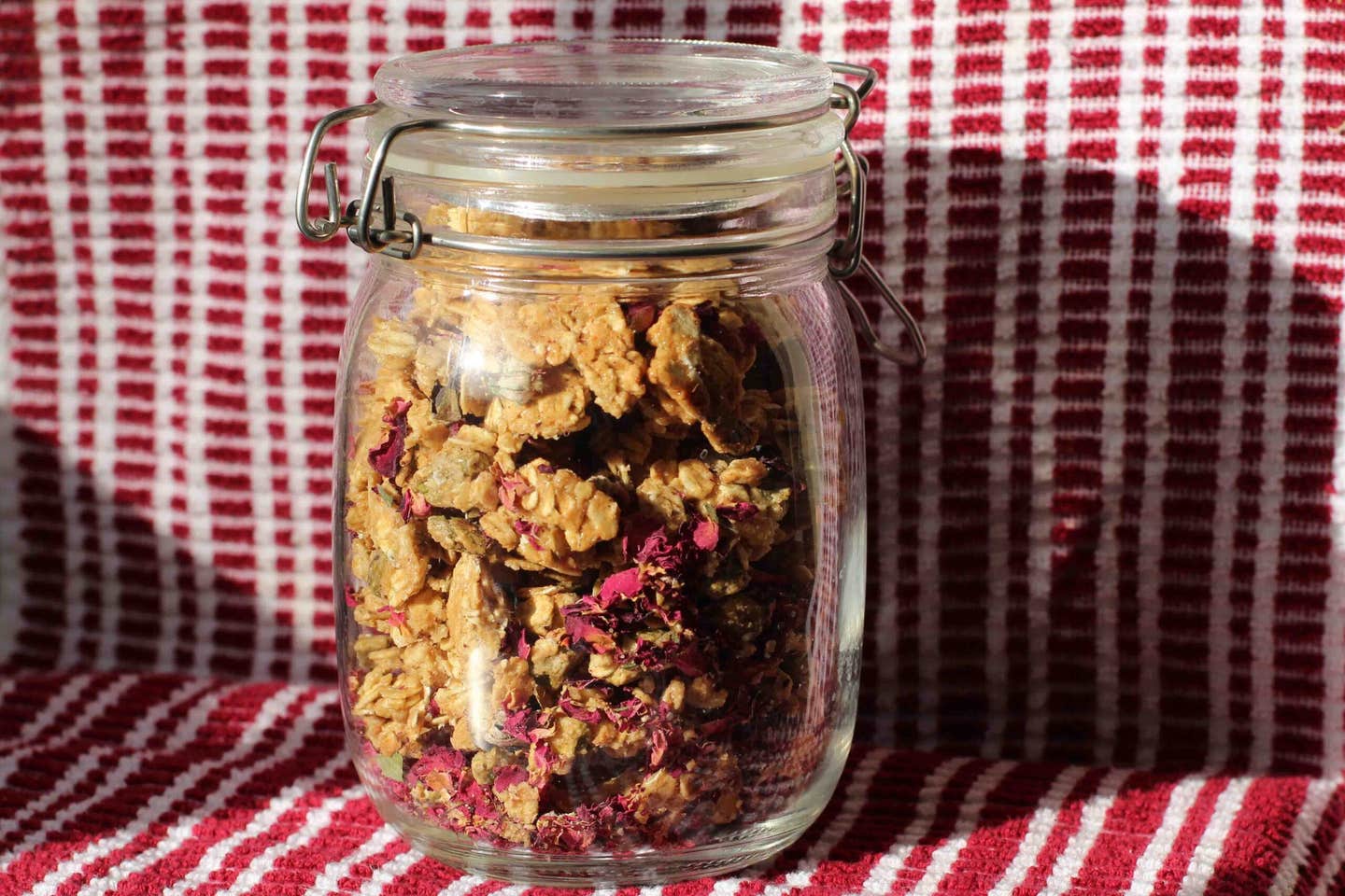 Rose Tahini Granola in a mason jar on a red and white striped tea towel underneath it and behind it