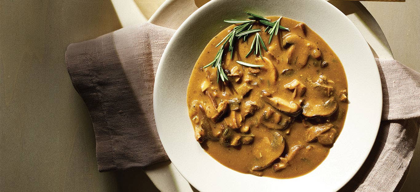 A creamy-looking bowl of Wild Mushroom Soup with a sprig of fresh rosemary on top. A brown cloth napkin sits under the white bowl.