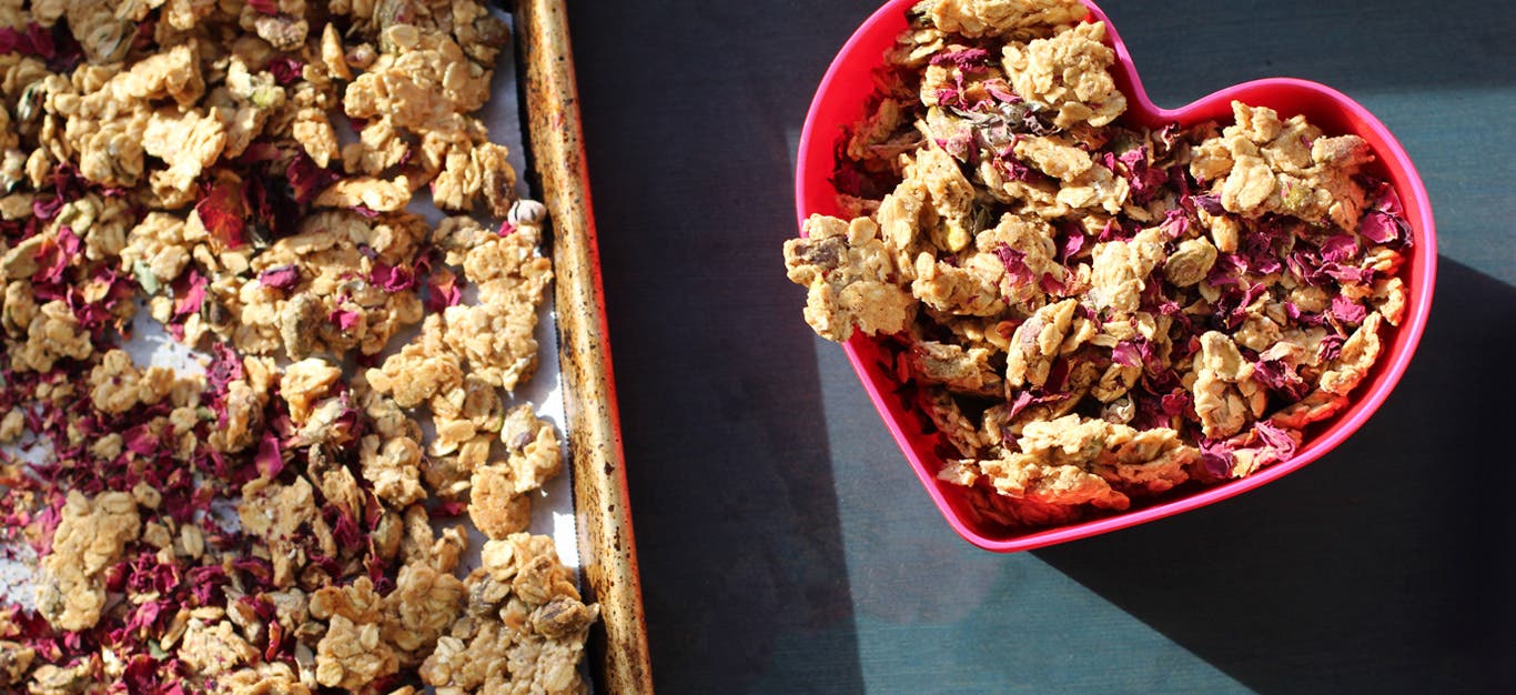 A ceramic baking dish filled with Rose Tahini Granola sits on a dark grey counter, next to a pink heart-shaped bowl of the granola