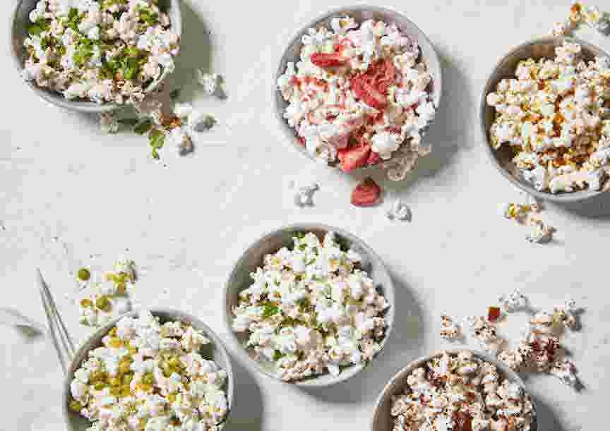 Six small bowls of oil-free popcorn, each topped with a creative homemade topper, including sweet and savory toppings