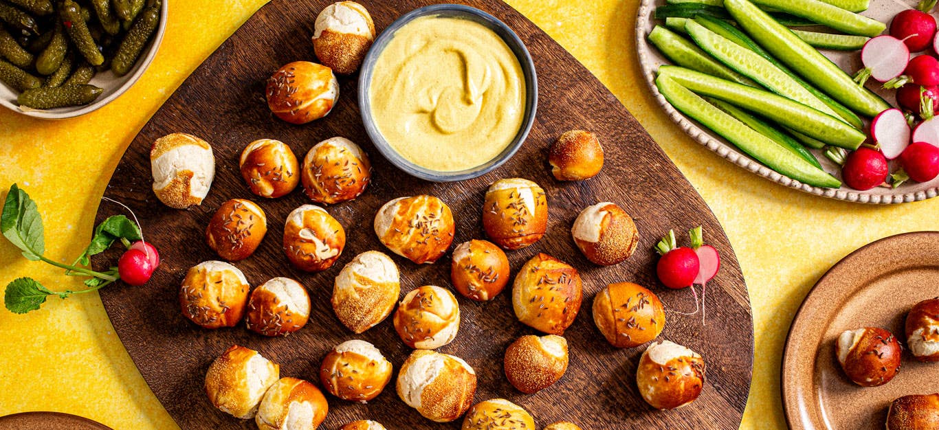 Whole Grain Pretzel Bites on a platter with mustard next to plates of sliced cucumber, radish, and pickles