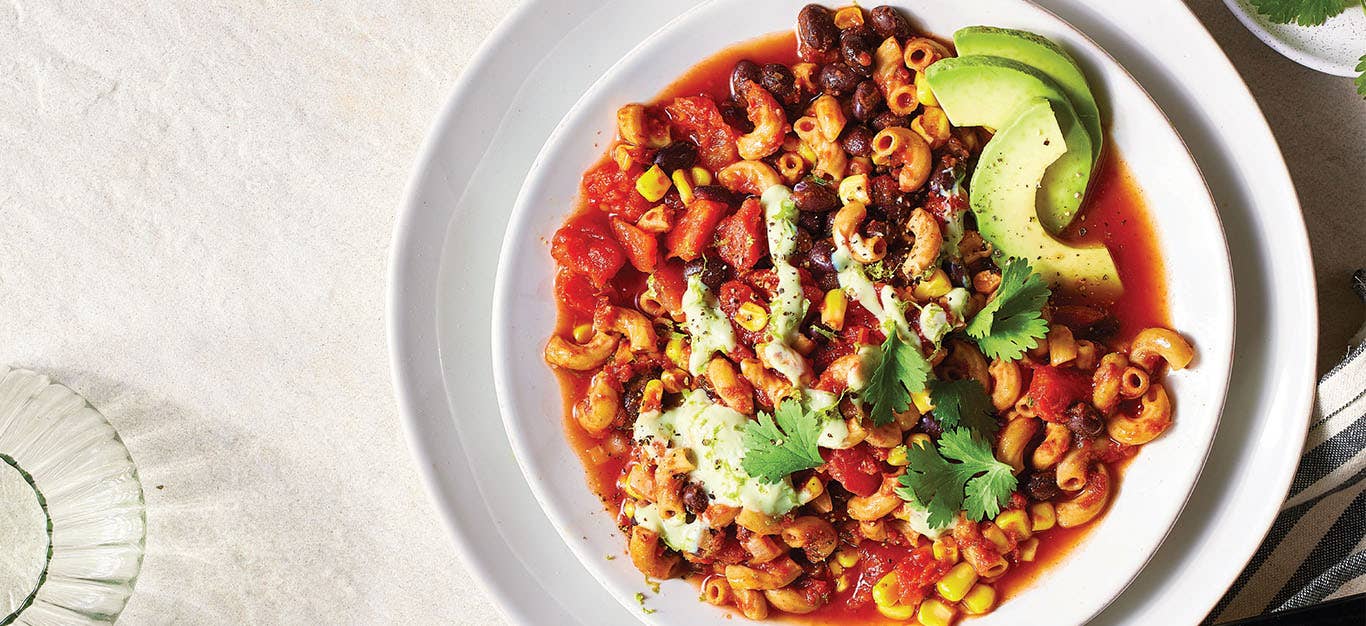 A white, wide-rimmed bowl of Zesty Macaroni with Black Beans, Corn, and Avocado Cream, nested on a white plate on a white table cloth