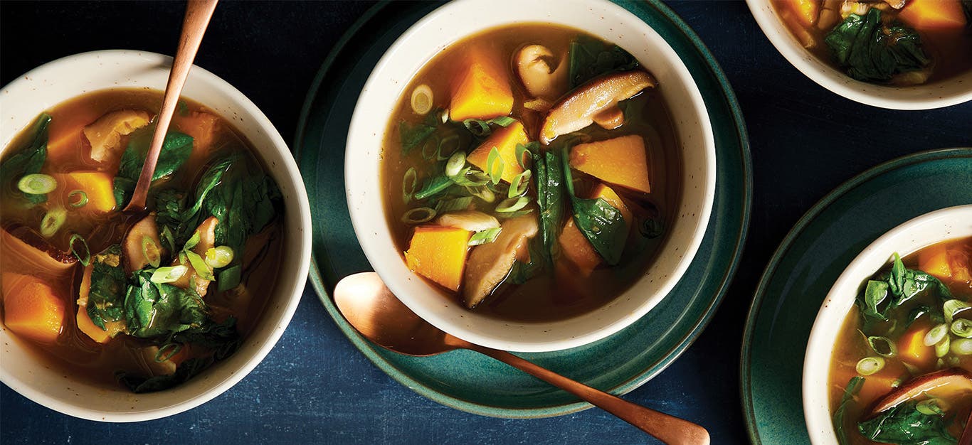 Four bowls of Thai Lemongrass Soup with Mushrooms, Squash, and Greens, on a dark blue tablecloth, with a spoon in each bowl