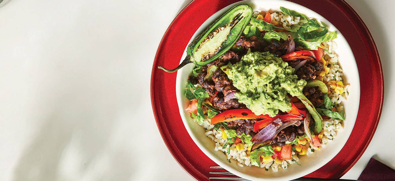 Veggie Burrito Bowl topped with guacamole, cooked red onion and red bell pepper, in a white bowl nested on a red plate