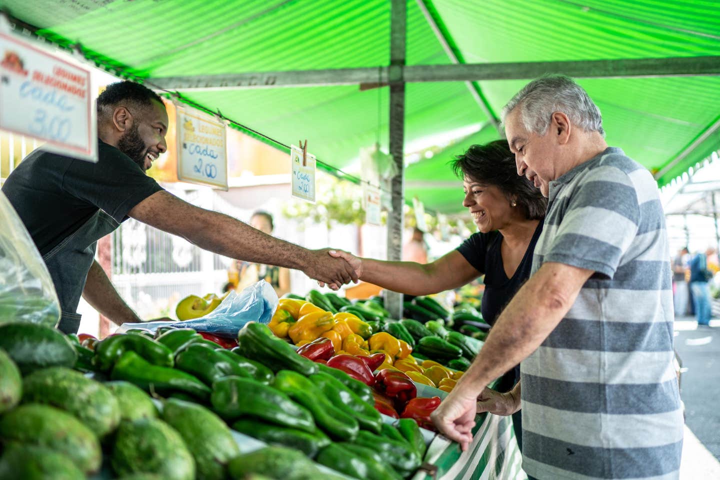 An older couple shops at a farmers market, looking at bell peppers, and the woman shakes the vendor's hand as they smile