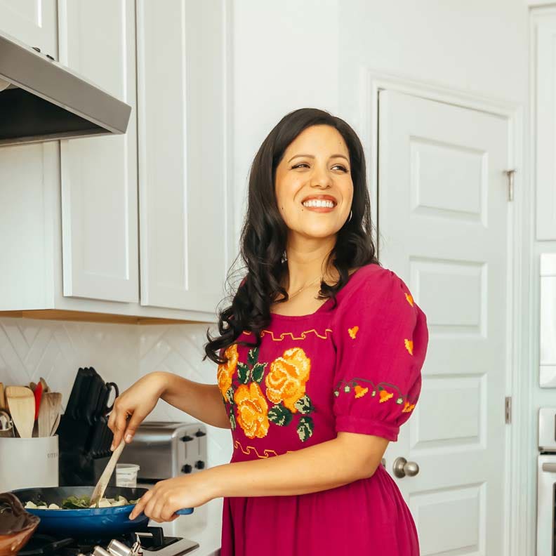 Chef and cookbook author Dora Ramírez stands in a bright kitchen over a skillet, wearing a magenta dress and smiling