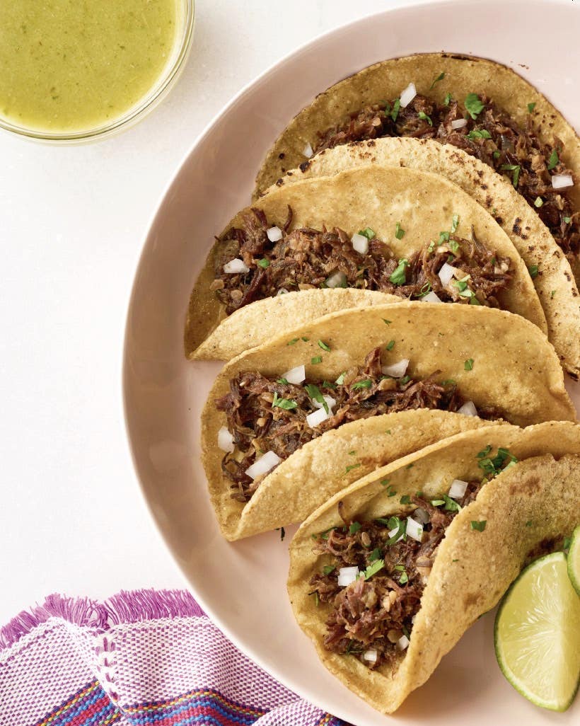 Hibiscus barbacoa tacos - four tacos filled with hibiscus flower barbacoa, a vegan barbacoa alternate, with two lime wedges on the side and a bowl of green sauce. Recipe from Comida Casera vegan Mexican cookbook