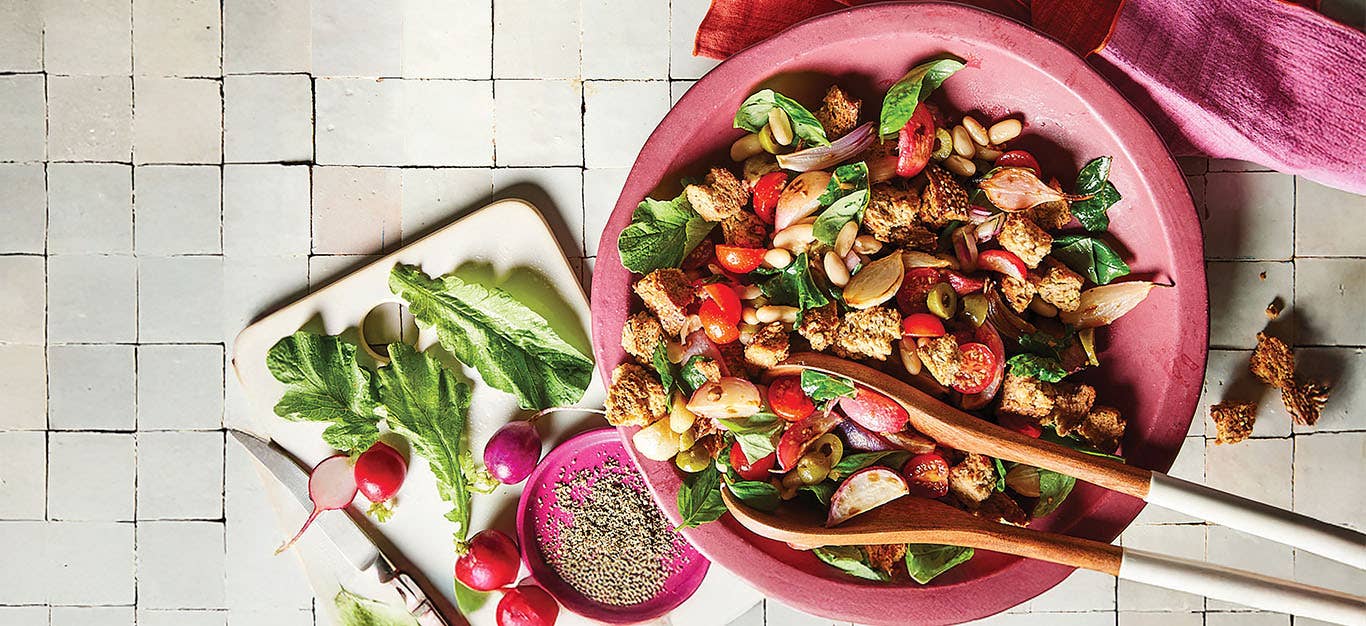 Easter Egg Radish Panzanella in a pink bowl on a white tiled background next to a plate of radishes and a small bowl of freshly ground pepper