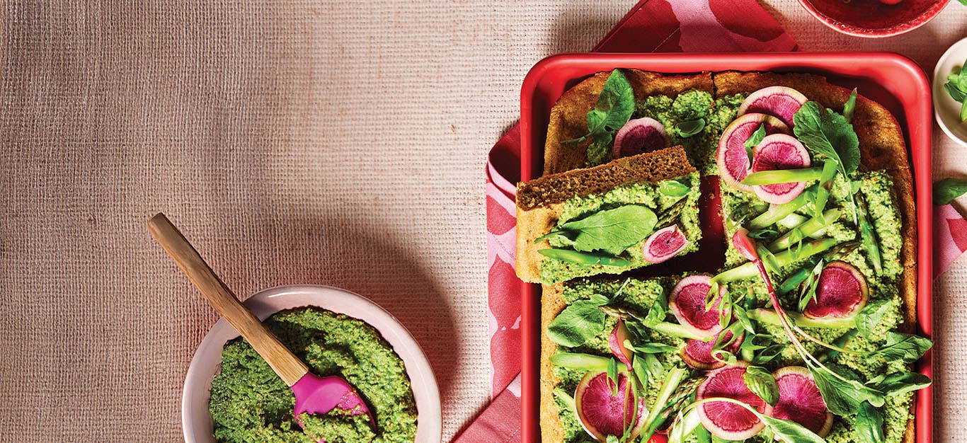 Spring Vegetable Focaccia in a red baking dish, topped witih colorful watermelon radishes and a herbaceous pea spread, next to a small bowl of the green spread