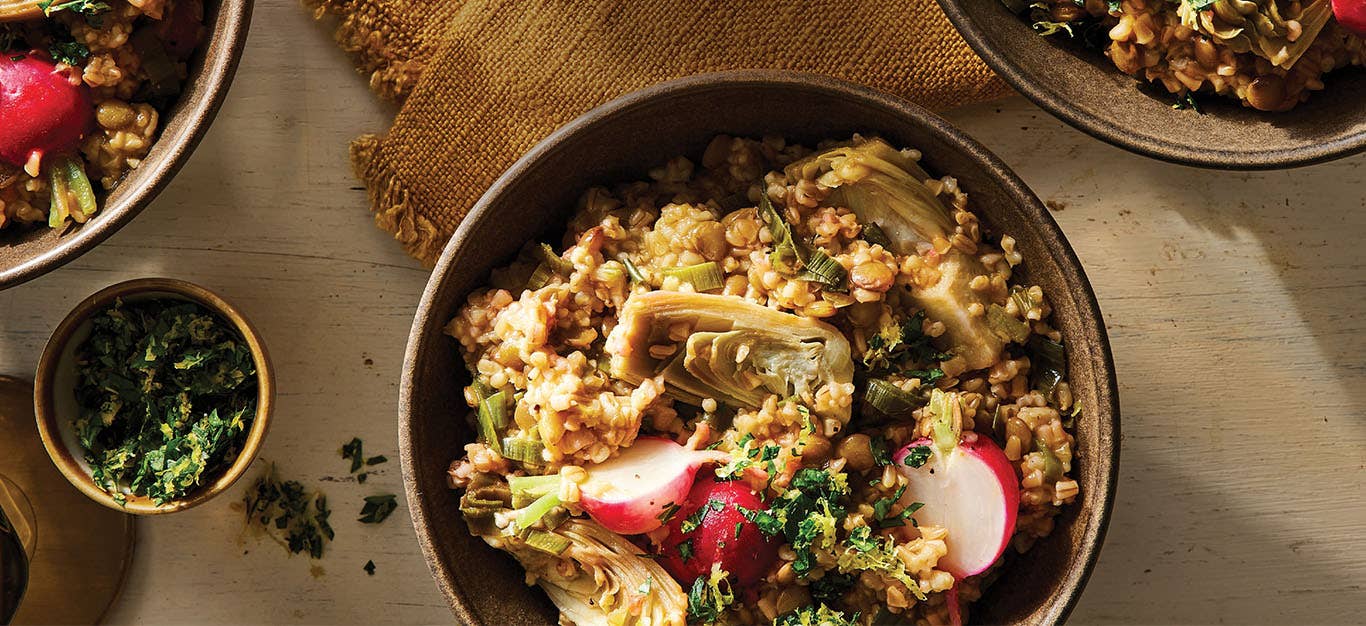 Steel-Cut Oat Risotto with Leeks and Artichokes in a brown ceramic bowl next to a small bowl of finely chopped parsley