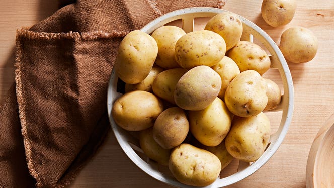 Around 20 new potatoes in a basket on a hardwood surface next to a rustic brown kitchen towel, with a couple new potatoes outsid the basket