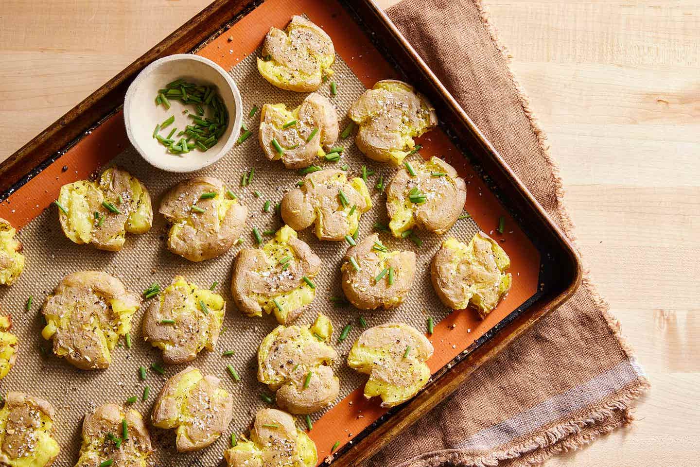 Crispy smashed new potatoes sprinkled with chives on a baking tray lined with a silpat mat. The tray sits on top of a rustic kitchen towel, which sits on top of a hardwood surface