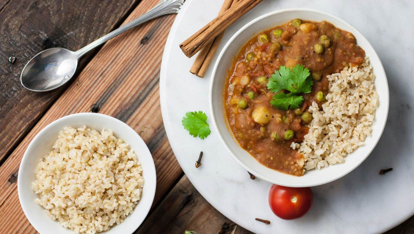 Aloo Mattar in a white bowl with brown rice on a marble chopping board with cinnamon stick and fresh tomatoes in the background