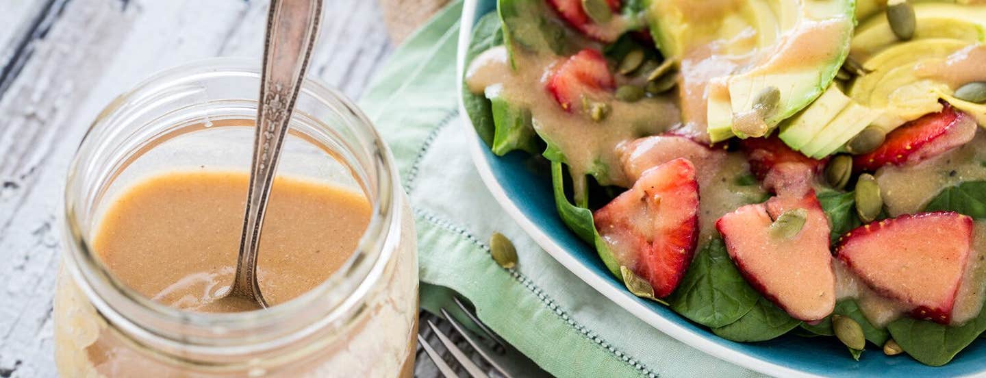 A mason jar of "Magical" Applesauce Vinaigrette next to a salad with fresh strawberries, spinach, and avocado, drizzled with the vinaigrette