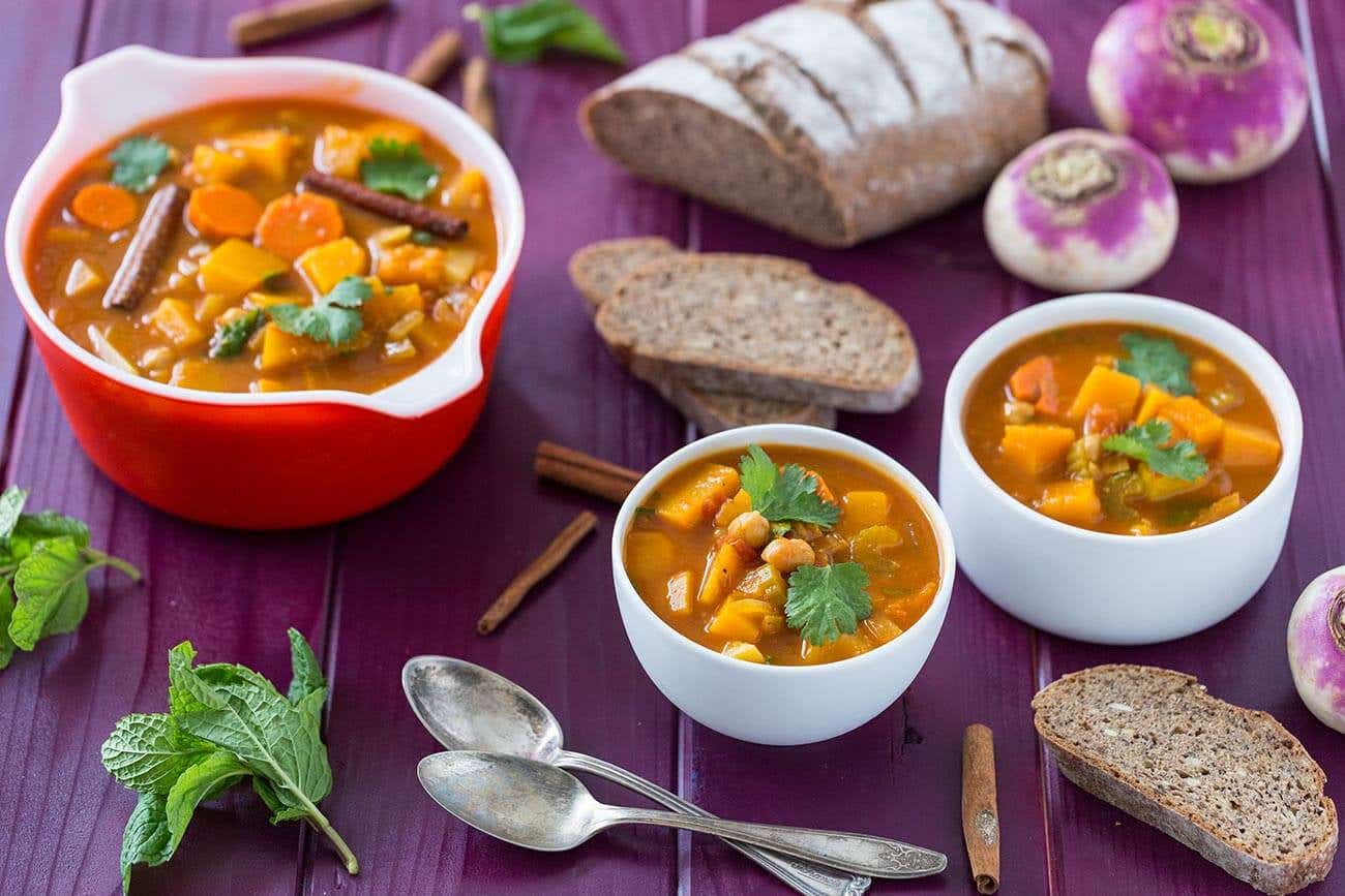 A large pot, and two smaller bowls of Vegetable Autumn Stew on a purple wooden table with chunks of whole grain bread and a couple of spoons