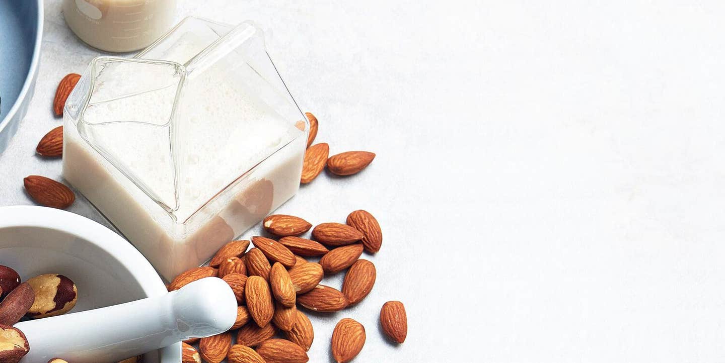 A glass carafe of nut milk next to whole almonds and a mortar and pestel