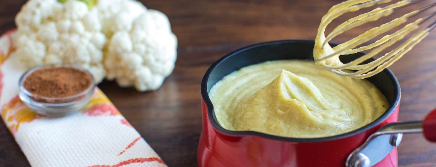 Cauliflower Bechamel in a small red pot, with a hand whisk held above the bowl and a fresh head of cauliflower in the background