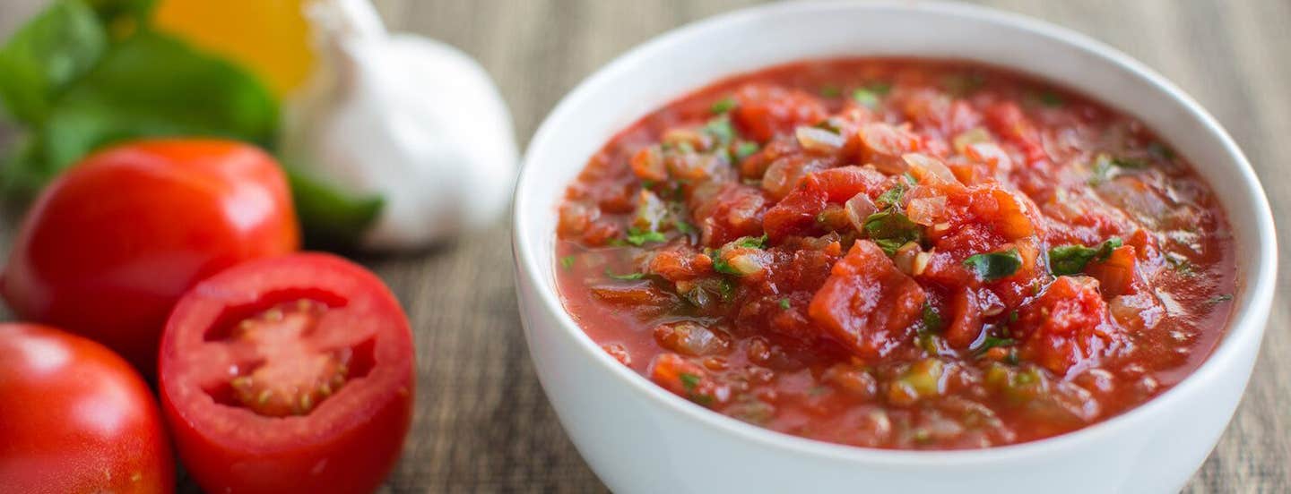 A bowl of tomato sauce with finely chopped fresh herbs to garnish, on a counter next to fresh tomatoes and a bulb of garlic