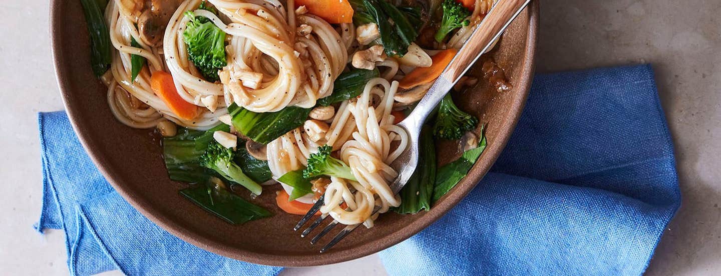 A wide-rimmed brown bowl of Chinese Noodles in Ginger Garlic Sauce next to a blue cloth napkin