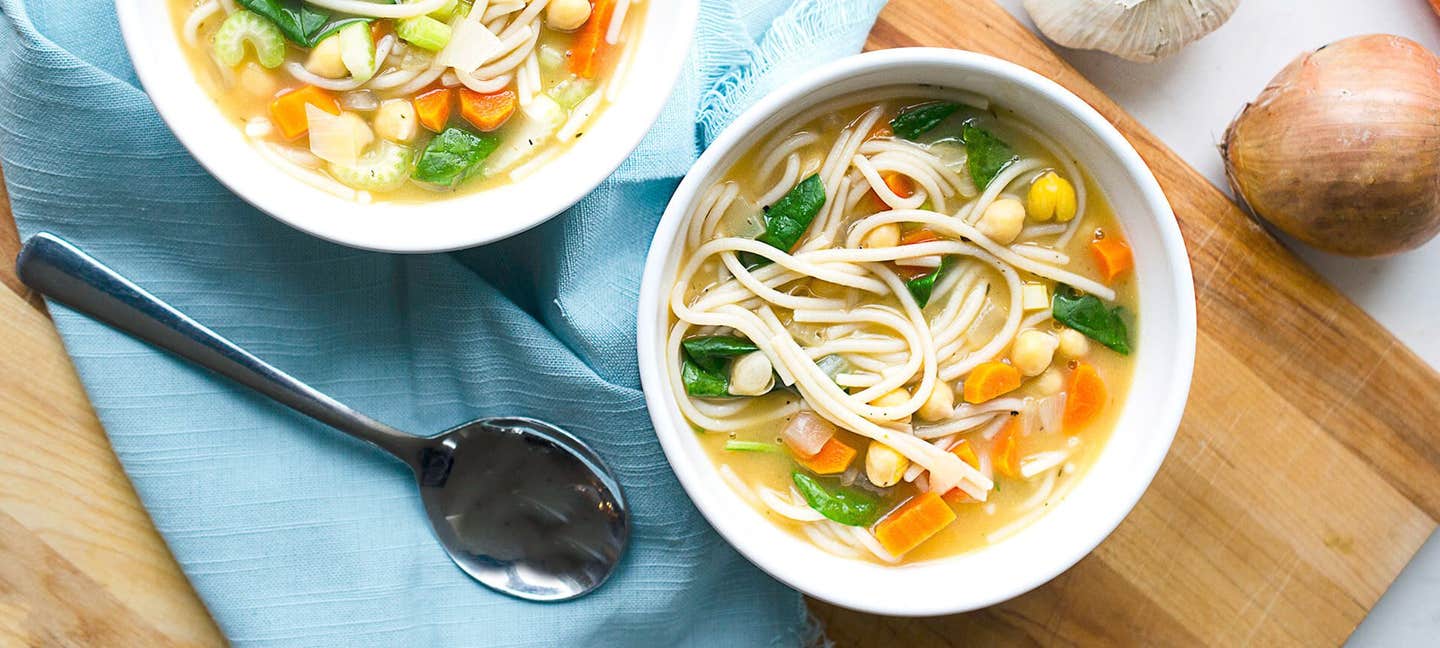 Two bowls of 30-Minute Chickpea Noodle Soup with Spinach with fresh onion and garlic to the side