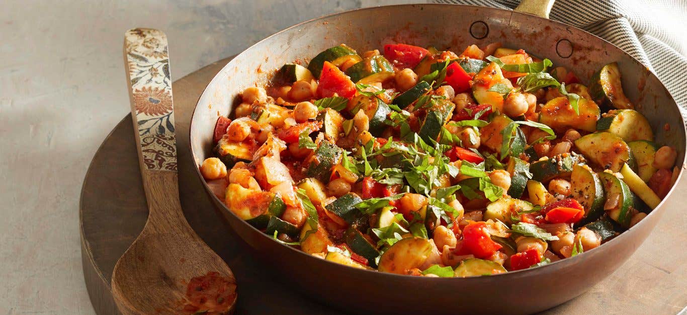 Chickpea Zucchini Saute in a large frying pan on a round wooden chopping board with a wooden serving spoon