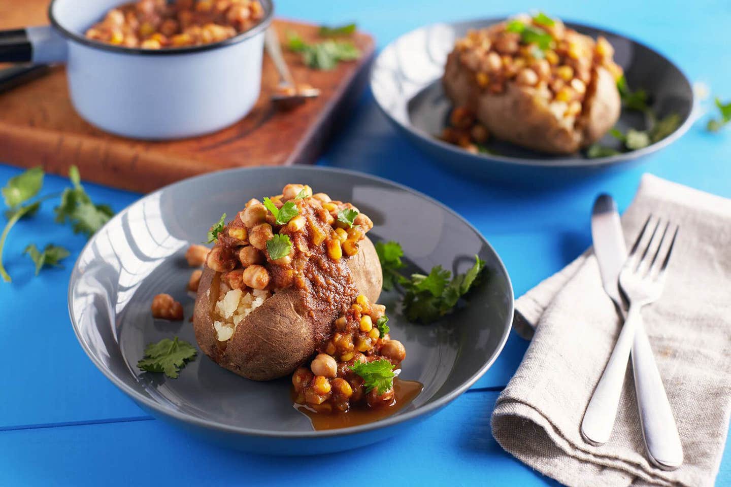 Two servings of Vegan Chickpea Chili on Baked Potatoes served in wide-mouthed bowls with a pot of Chickpea Chili in the background