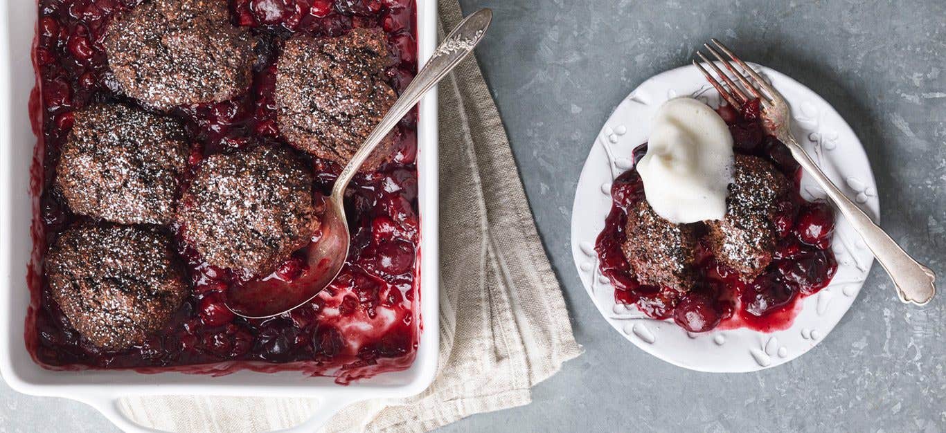 Chocolate Cherry Cobbler shown in a baking dish with serving spoon, with one serving on a plate shown with whipped cream topping