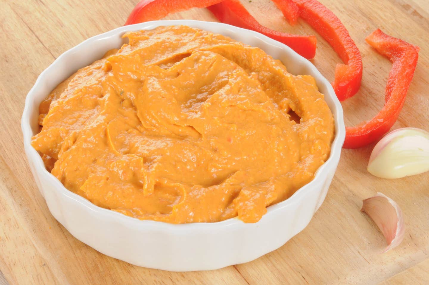 Roasted Red Pepper Tapenade Hummus in a white bowl next to fresh garlic cloves a slices of red bell pepper