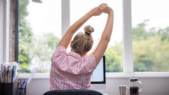 Woman in a pink shirt stretching arms overhead while seated at a desk