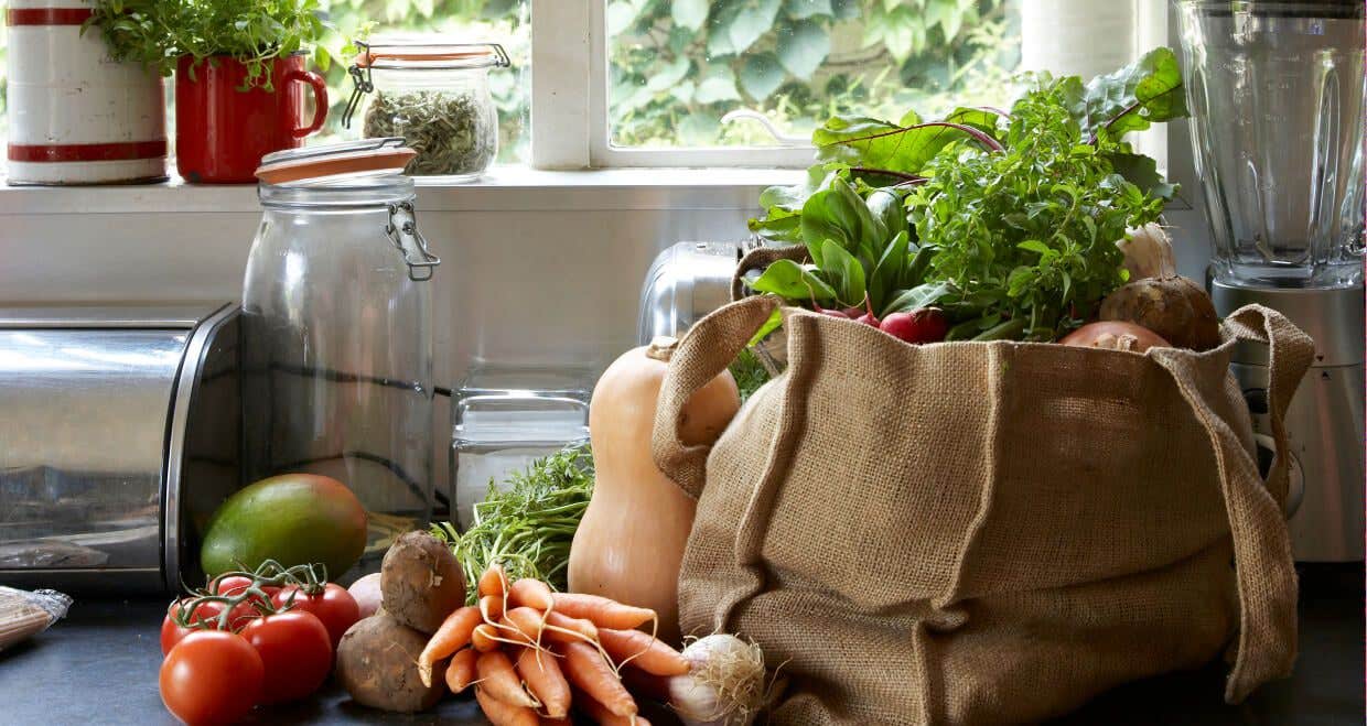 A cloth bag filled with fresh veggies on the kitchen counter next jars, a blender, more veggies