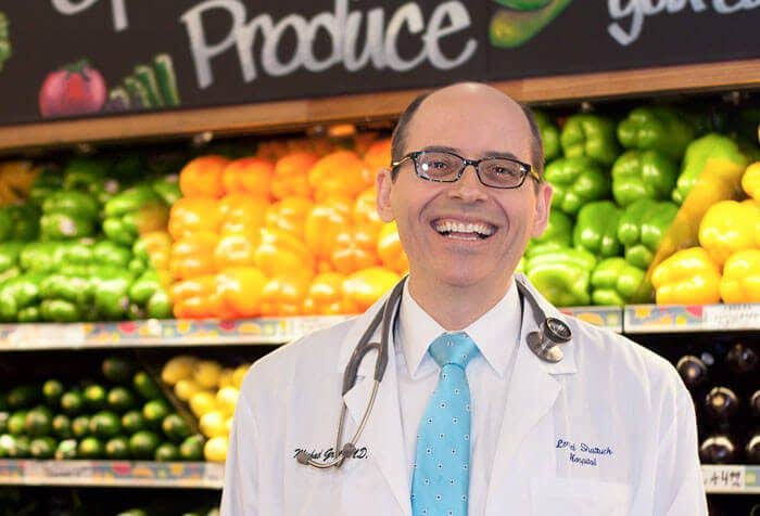 A laughing Dr Michael Greger in a white lab coat at the veggie section of the supermarket