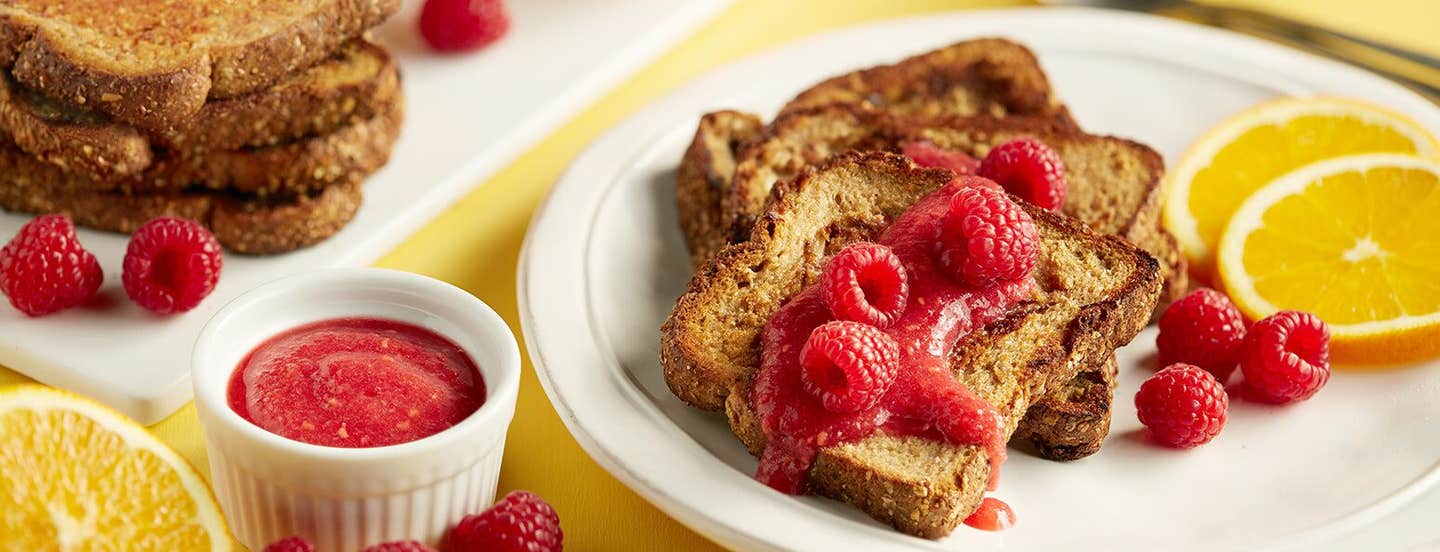 On a bright yellow table is a white plate with vegan Orange French Toast, fresh raspberries, orange slices, and a raspberry compote. Next to the plate is a small white ramekin of extra compote.