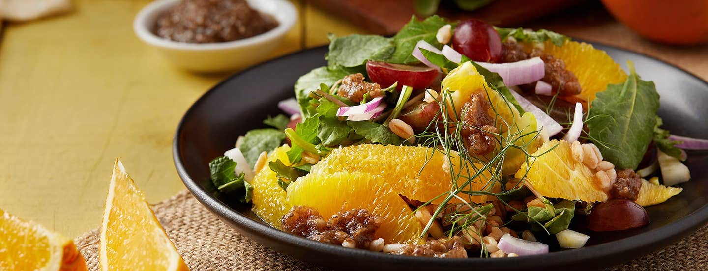 Baby Kale Salad with Farro, Grapes, and Oranges in a black serving bowl with wedges of orange to the sice