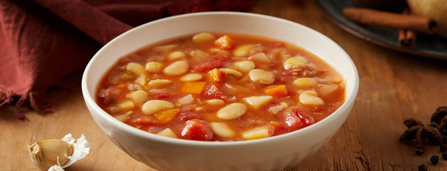 Autumn Lima Bean Stew in a white bowl on a rough wood table with a plate of potato, onion, and cinnamon stick in the background