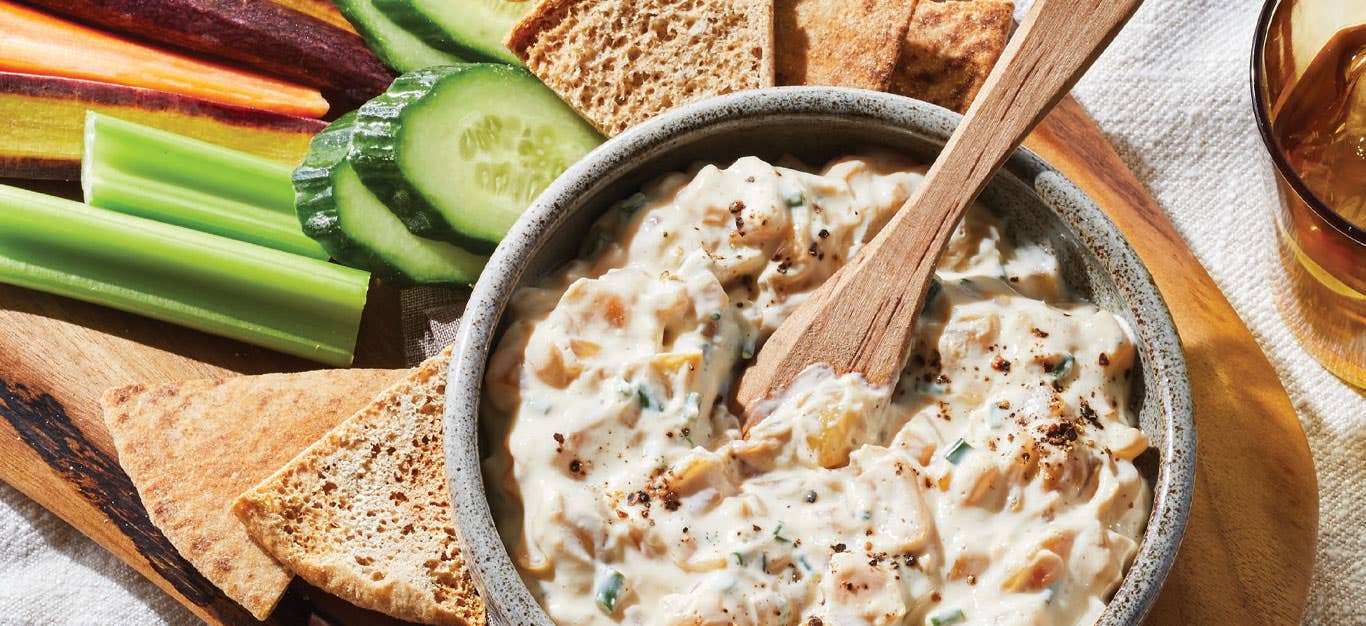 Vegan French Onion Dip in a gray bowl with a wooden spoon on a serving board with pita chips and sliced veggies