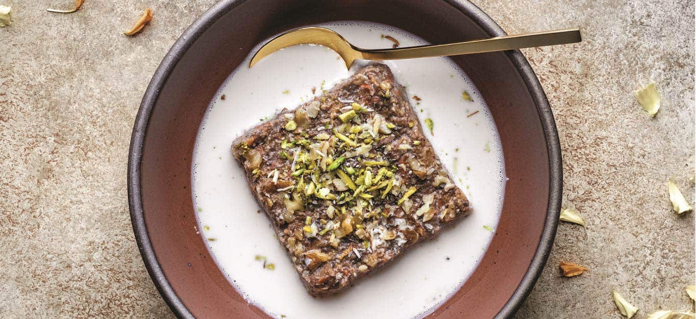 Gajjar No Halvo Baked Oatmeal in a brown bowl, with white milk surrounding the oatmeal square; a garnish of flower petals is visible around outside of the bowl