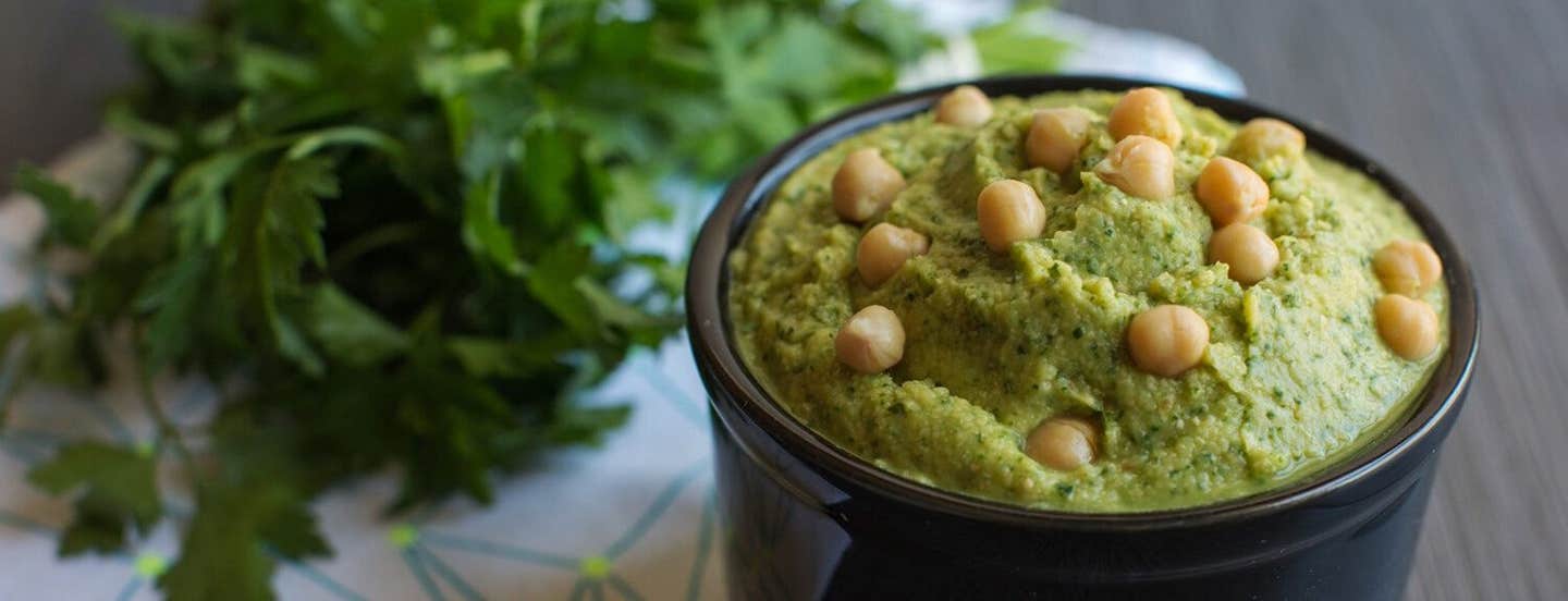 Herbed Hummus in a small black serving bowl, garnished with whole chickpeas and with a bunch of flat-leaf parsley in the background