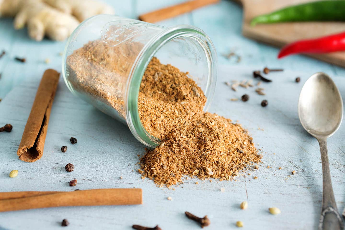 A small mason jar on it's side on a pale blue wooden counter, with a seasoning spilling out. Around the jar are cinnamon sticks, fresh ginger, cloves, and fresh chiles.