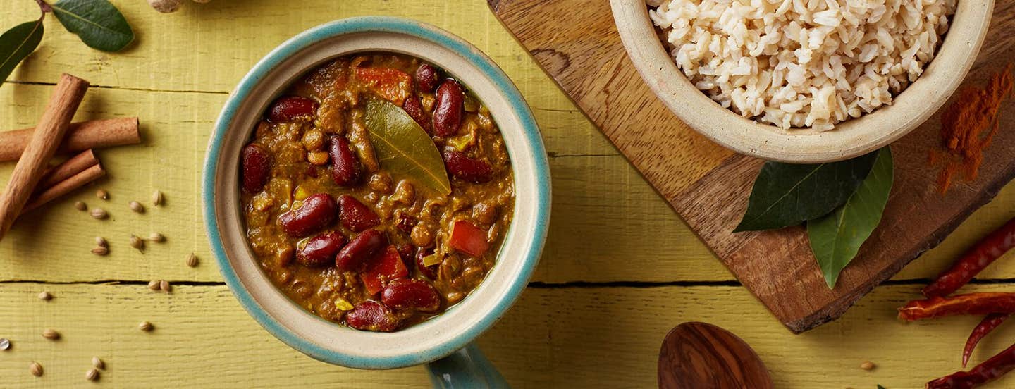 Top down view of Kidney Bean and Lentil Dal on a mustard counter next to a wooden chopping board, a bowl of brown rice, and cinnamon sticks