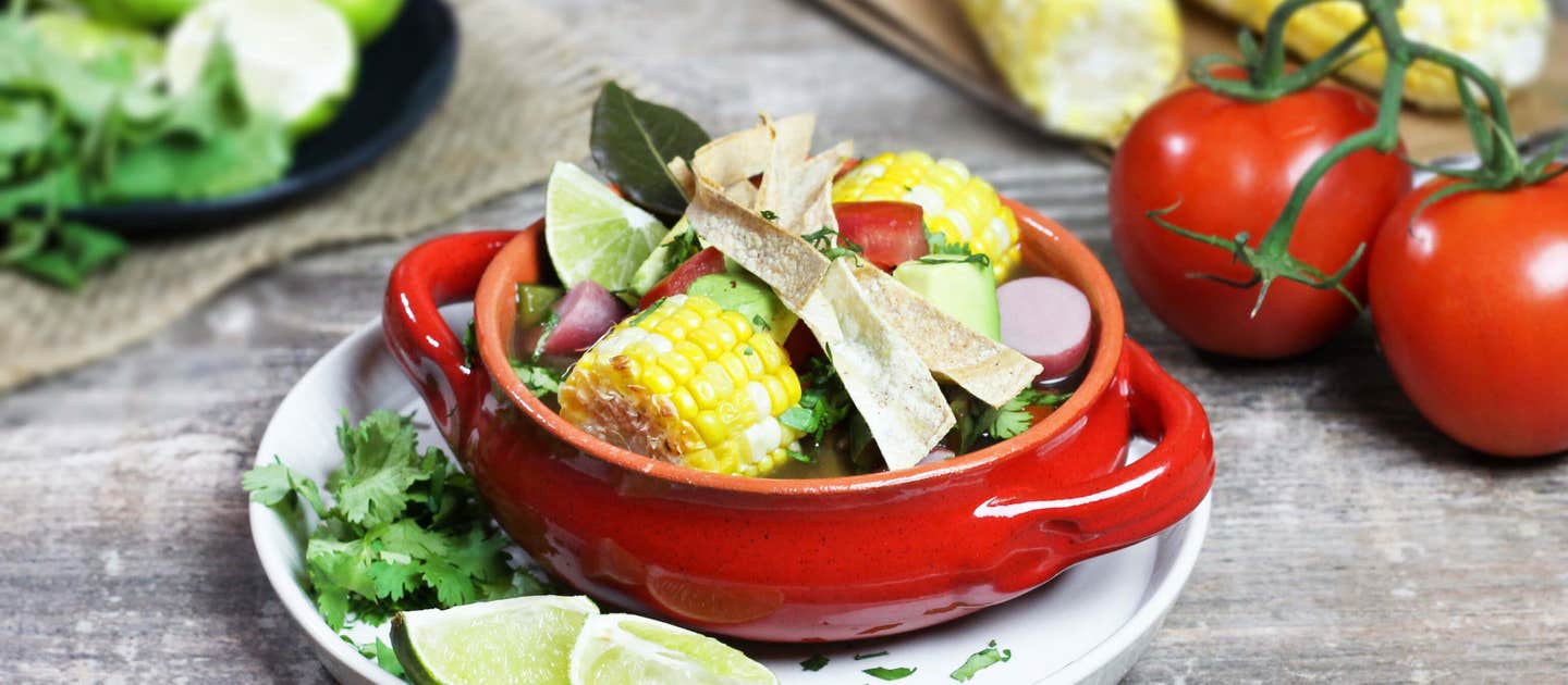 Mexican Lime Soup in a white two-handled soup bowl on a plate with lime wedges, fresh cilantro, and with fresh strawberries and corn in the background