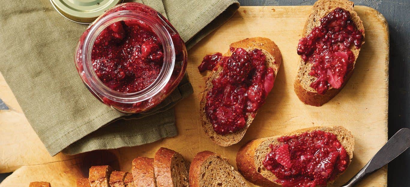 Homemade mixed blackberry, raspberry, and strawberry jam spread on a few slices of whole grain bread with a jar of it off to the side