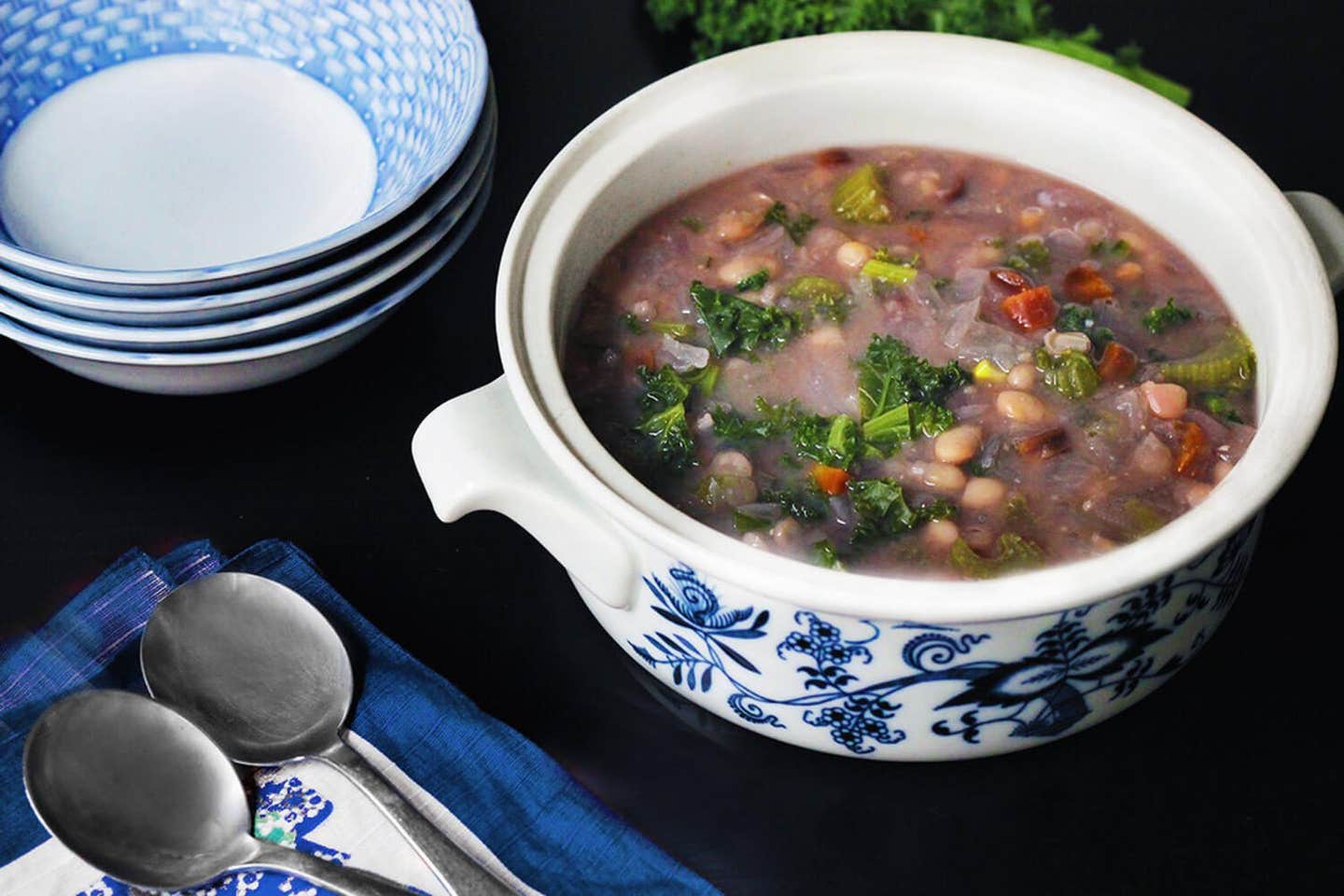 A large white pot of Navy Bean Soup next to a stack of blue and white bowls and a couple of spoons