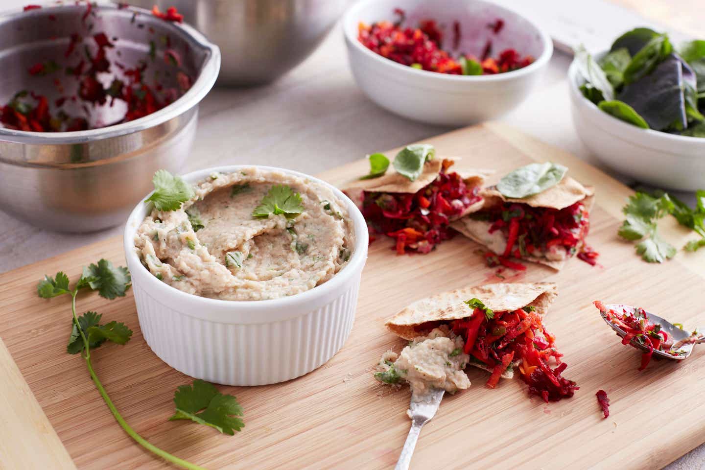 A ramekin of white navy bean hummus sprinkled with fresh herbs is on a cutting board beside whole wheat pitas and fresh veggies